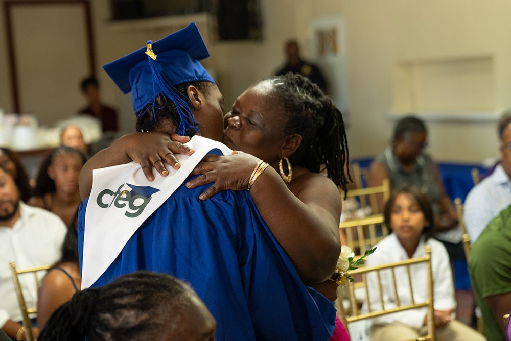 A CFEG graduate in a blue cap and gown, wearing a white CFEG stole, embracing a family member at commencement.